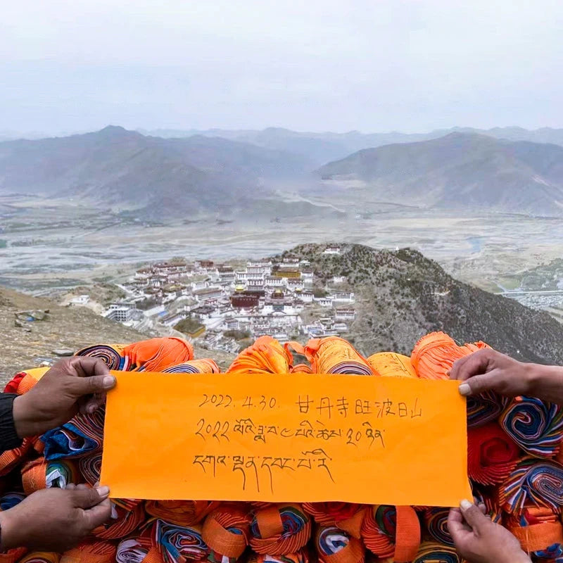 Hilltop monastery and wide mountain landscape with hands holding an orange note above stacked tibetan flags for prayers of blessing.