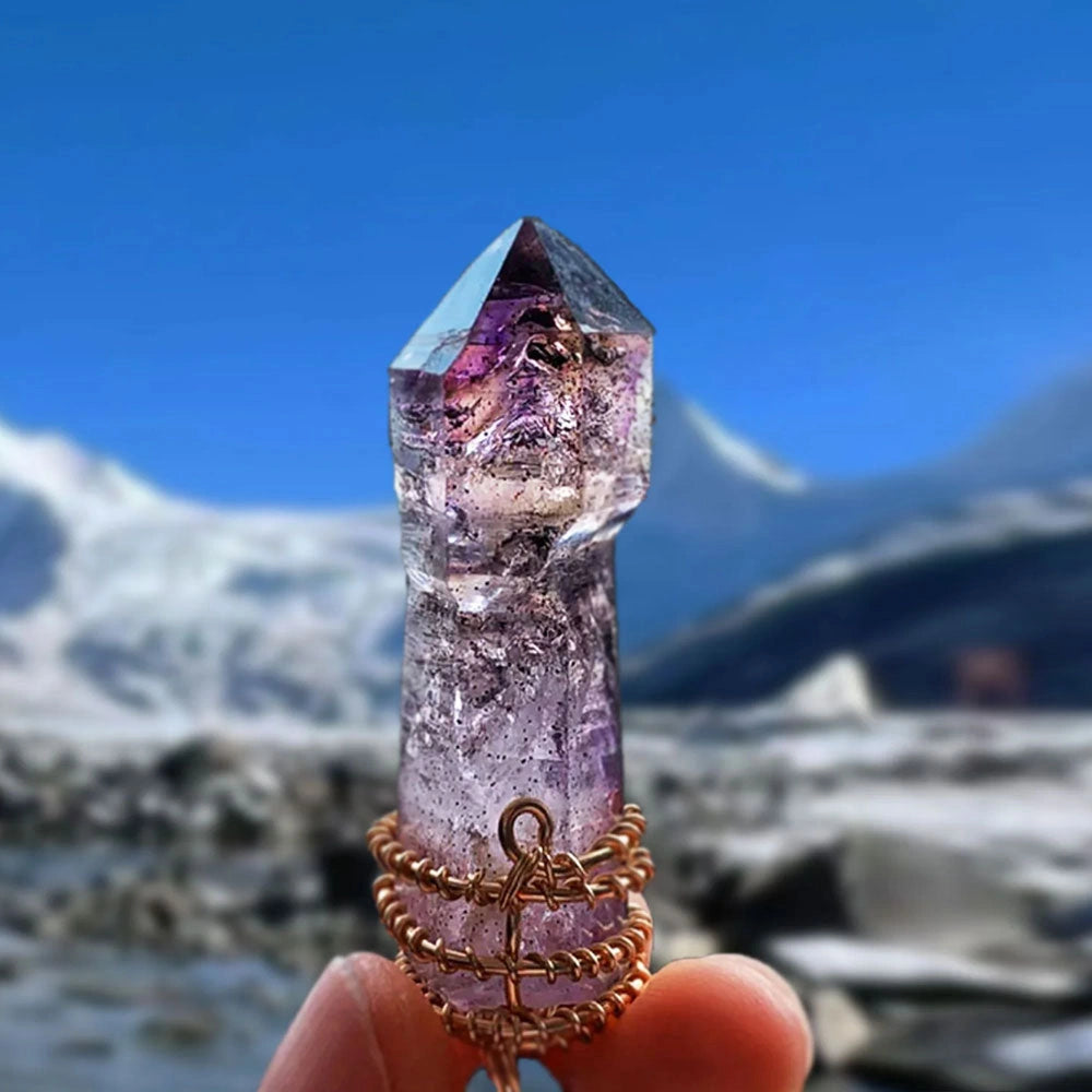Hand holding a wire-wrapped purple quartz point with snowy mountains blurred in the background.
