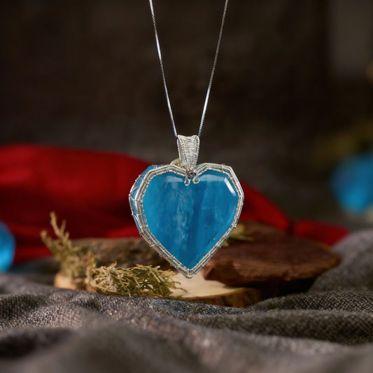 Blue heart-shaped pendant on a chain resting on fabric with a wooden slab and red cloth behind.
