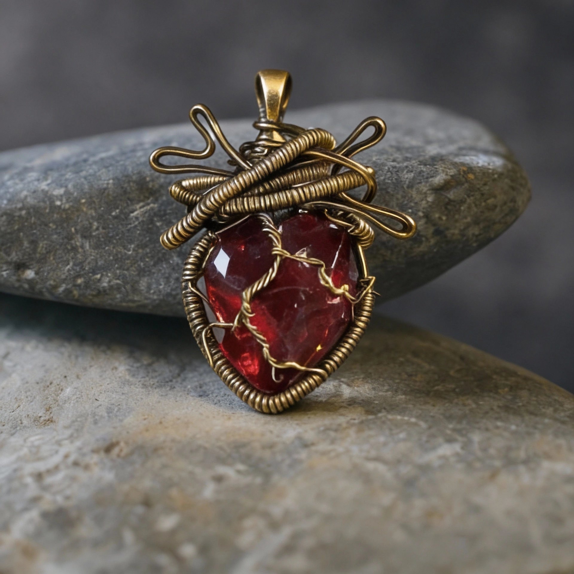 Red stone pendant in intricate brass wire wrap displayed on smooth gray river stones.