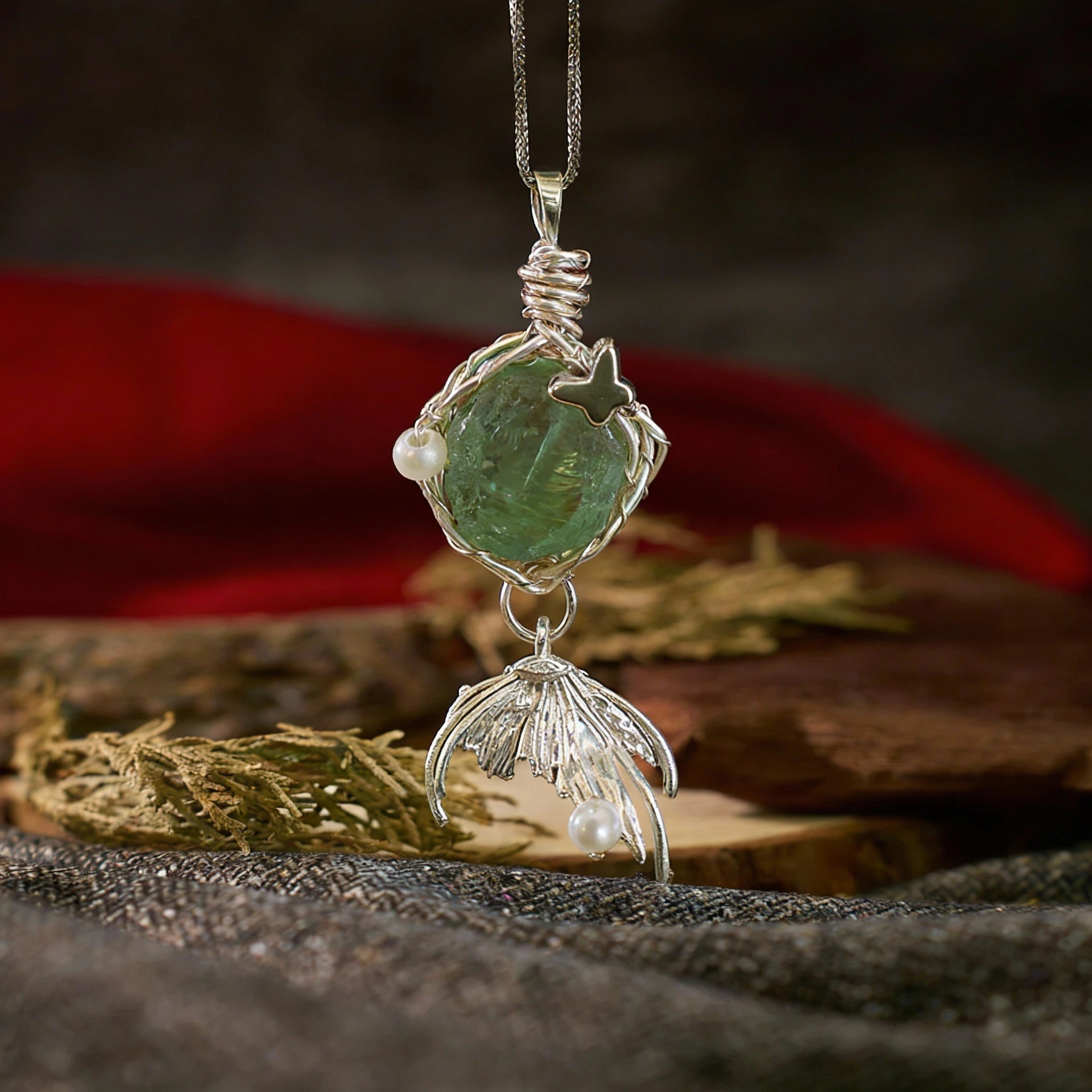 Green rough-cut stone pendant with silver leaf-like dangle, displayed on fabric with red cloth and wooden slab.