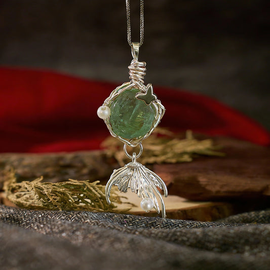 Green rough-cut stone pendant with silver leaf-like dangle, displayed on fabric with red cloth and wooden slab.