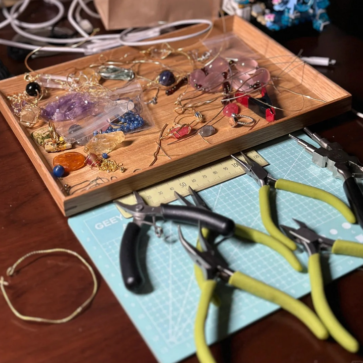 Workbench for handmade crystal pendant and wire‑wrapped necklace making—pliers and tools on a cutting mat beside trays of heart-shaped gemstones, beads, and chains for boho artisan jewelry and talisman amulets.