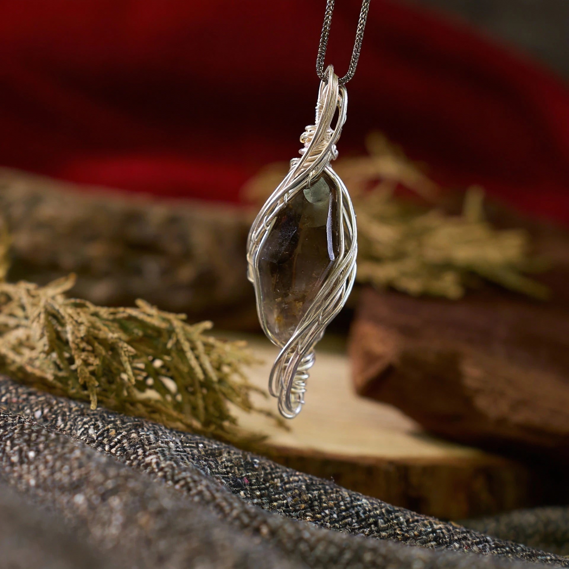 Smoky translucent stone wrapped in twisting silver wires, displayed on fabric with dried plants and wood.