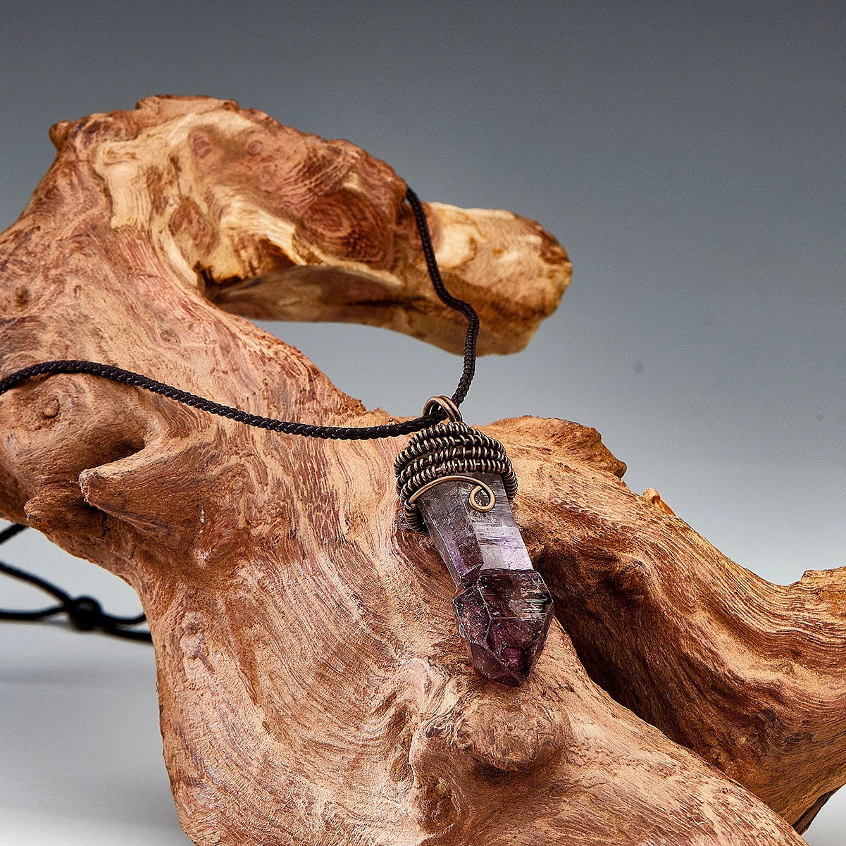 Small amethyst pendant on a dark cord displayed on a gnarled wood prop.