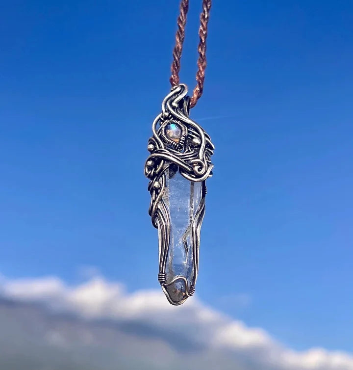 Silver-wrapped crystal pendant on a braided cord against a blue sky.